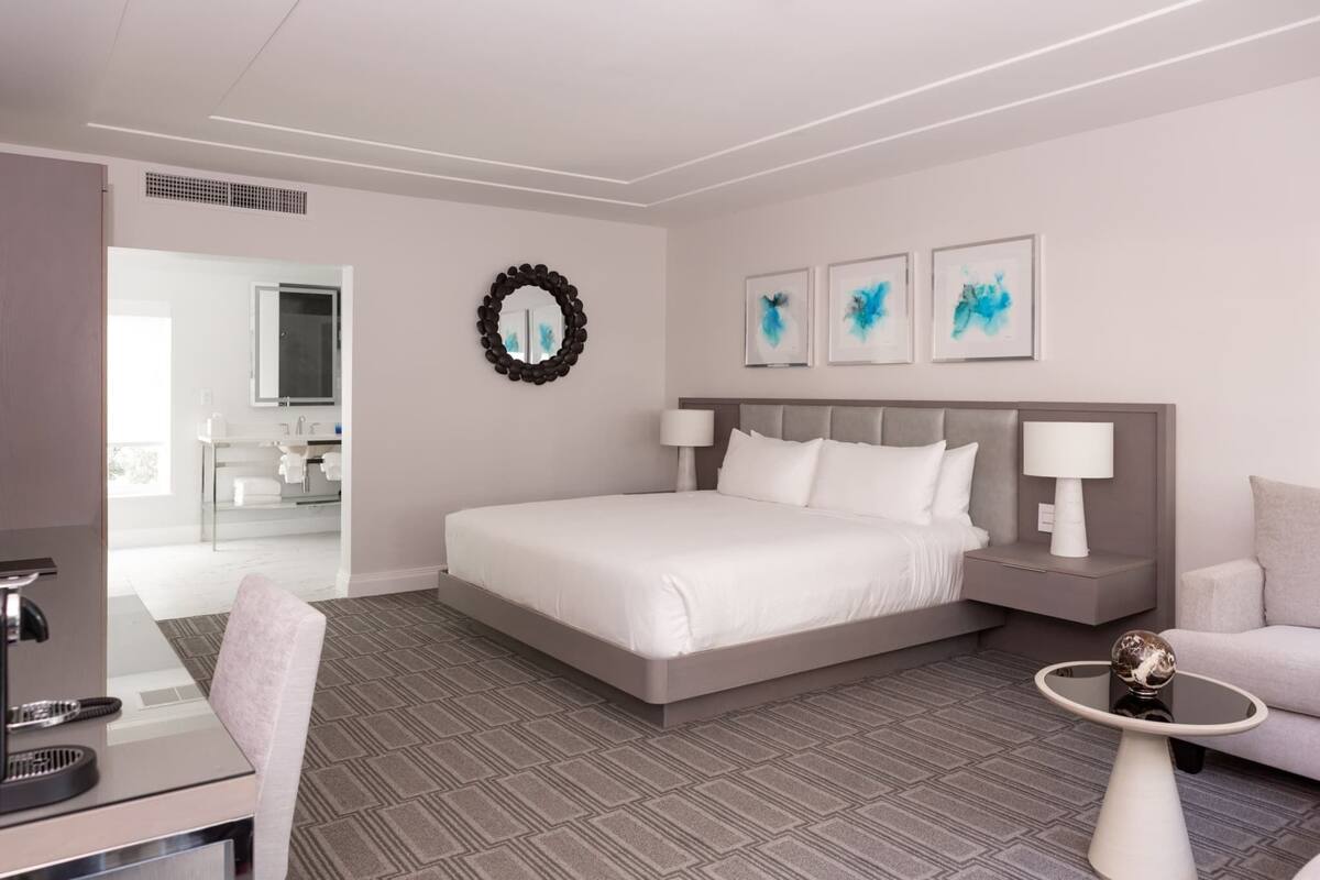A hotel room with a bed, bedside tables, a desk, a chair, and artwork on the wall.