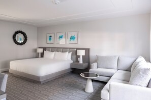 Una habitación de hotel moderna con una cama grande, un sofá gris, una mesita redonda y un espejo montado en la pared.