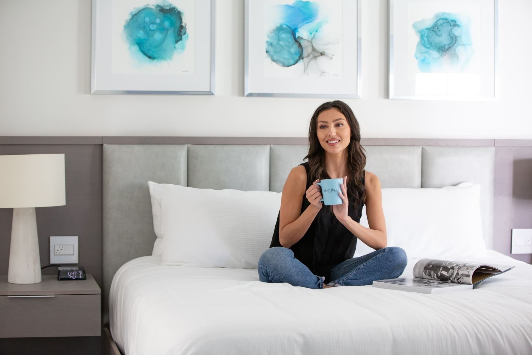 A person sitting on a bed with a digital clock, a magazine, and a blue mug.