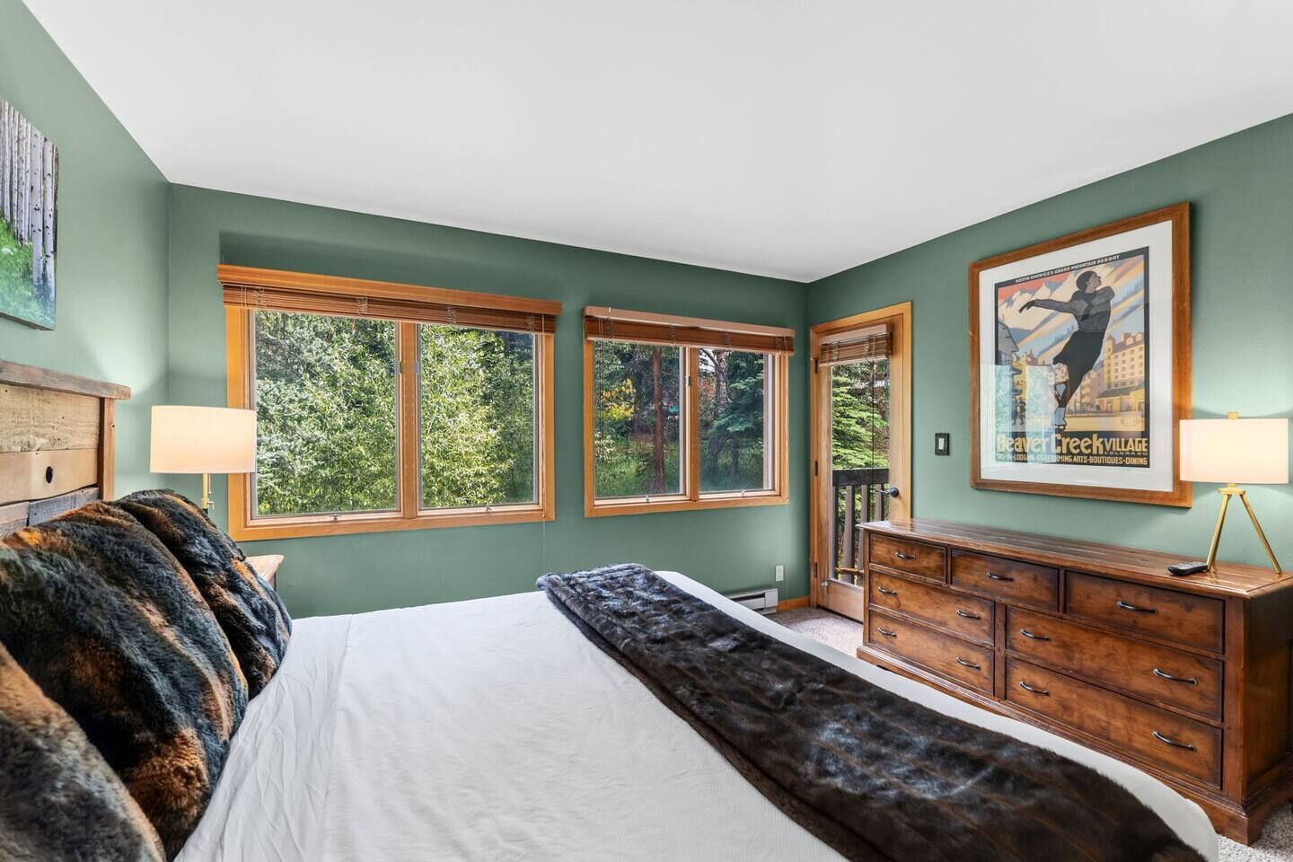 A bedroom with a large bed, wooden dresser, and two windows with a view of trees.