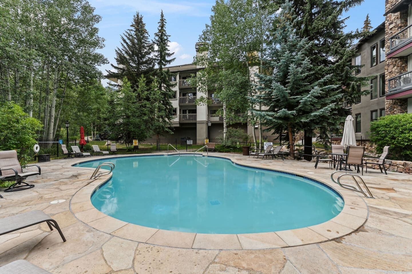 A swimming pool with lounge chairs and umbrellas, surrounded by trees and a multi-story building.