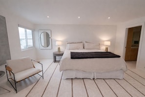 Ein modernes Schlafzimmer mit einem großen Bett, Nachttischlampen, einem Sessel und Blick ins Freie.
