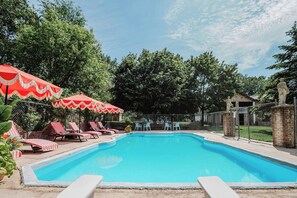 Piscina com espreguiçadeiras e guarda-sóis, rodeada por árvores e um muro de tijolos.