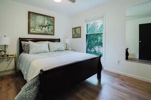 Quarto com cama de madeira, duas luminárias de cabeceira, uma janela grande com vista para o verde e duas molduras na parede.