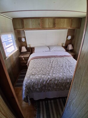 A bedroom with a wooden headboard, a bed with floral bedding, two bedside lamps, and a window with blinds.
