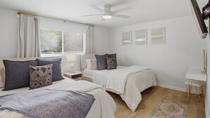 Ein Schlafzimmer mit zwei Betten, einem Deckenventilator, einem Fenster mit Jalousien, einem Nachttisch mit Lampe und einem an der Wand befestigten Fernseher.