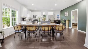 Una cocina amplia con armarios blancos, un comedor con mesa de madera y sillas de mimbre, y vista a la vegetación a través de ventanas grandes.