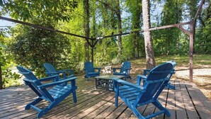 Terraza de madera con sillas Adirondack azules y una pequeña mesa, rodeada de árboles.