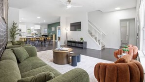 Una sala de estar moderna con un sofá, una mesa de centro y un reposapiés, un comedor con sillas y una mesa, una cocina al fondo, y una escalera.