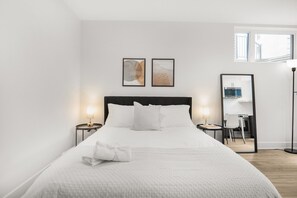 A hotel room with a large bed, two bedside tables with lamps, a desk with a chair, and a mirror. - Stylish Queen Studio Apartment with Fast Wi-Fi & Parking (Ottawa)