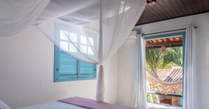 A bedroom with a bed, a window with blue shutters, and a view of a tiled roof outside.