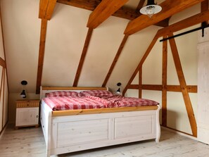 A bedroom with a sloped ceiling, wooden beams, a white bed with a checkered blanket, two bedside tables, and a hanging light fixture. - Experience monument protection: Stylish half-timbered attic with a feel-good climate (Kottmar OT Obercunnersdorf)