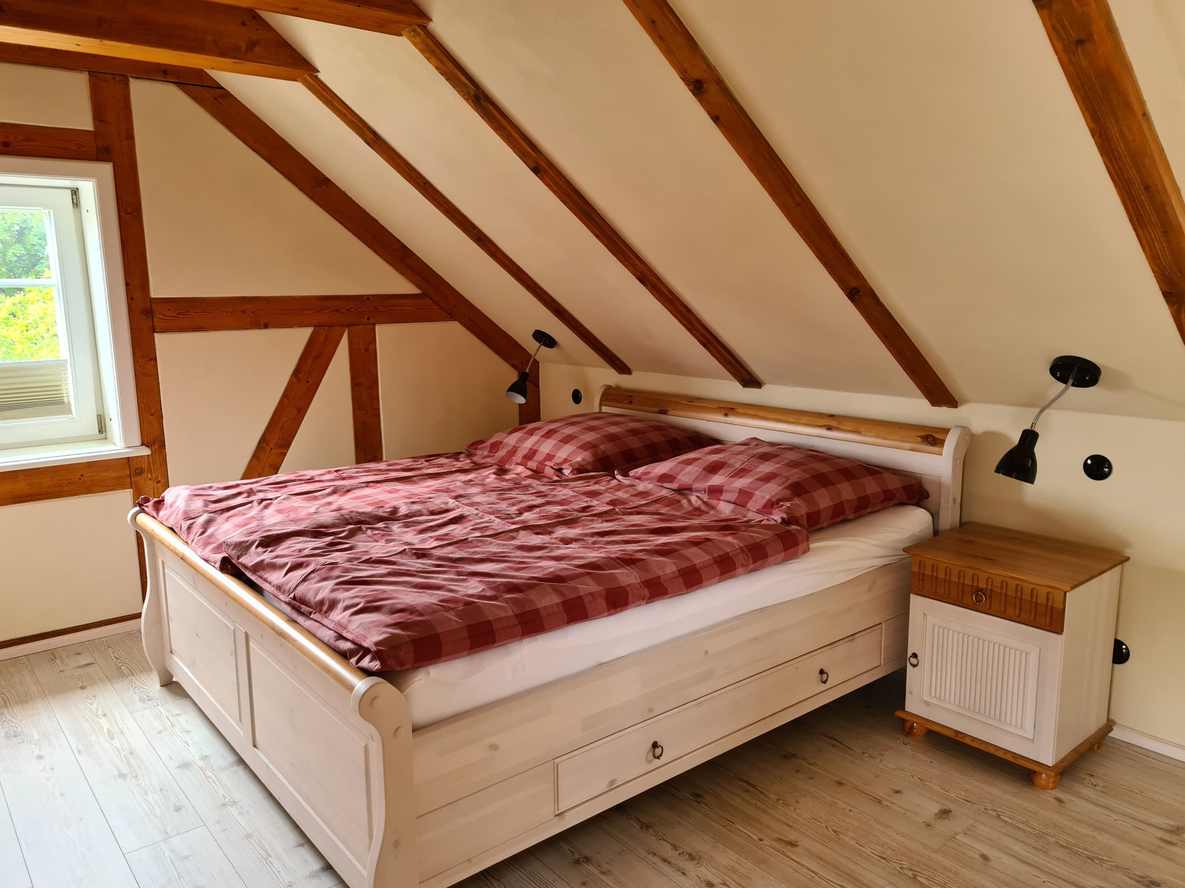 A bedroom with a wooden ceiling, a bed with a checkered blanket, a bedside table, and a window.