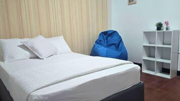 A bed with white bedding, a blue bean bag chair, and a white shelving unit with a plant.