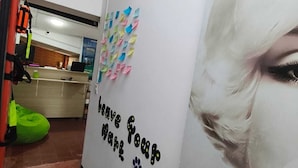 Un pasillo con una pared blanca que muestra un letrero que dice 'Traveler's Wall' y un cartel con un rostro borroso. Hay notas adhesivas de colores en la pared y un sillón de pie verde en el fondo.