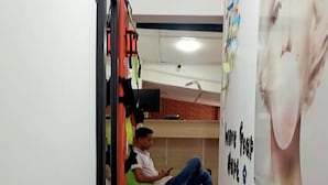 Un pasillo estrecho con suelo de baldosas, una persona sentada en un sillón de mimbre, y una pared con un cartel.