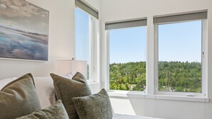 Ein Schlafzimmer mit einem großen Bett, vielen Kissen, einem Gemälde an der Wand und Blick auf einen Wald durch zwei Fenster.