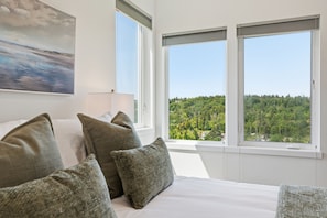 Ein Schlafzimmer mit einem großen Bett, vielen Kissen, einem Gemälde an der Wand und Blick auf einen Wald durch zwei Fenster.