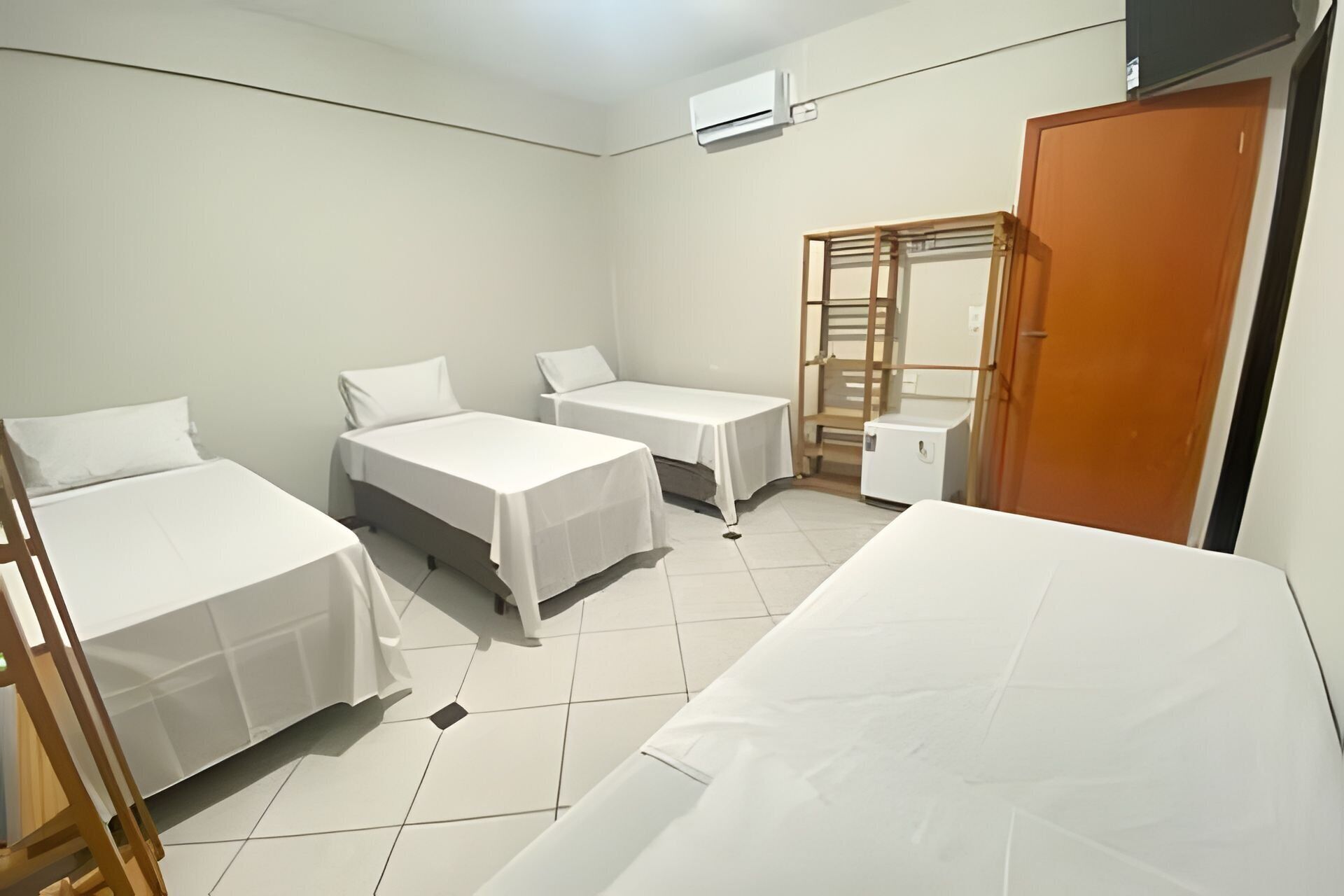 A hotel room with three beds, a wooden wardrobe, a small table, and an air conditioning unit.