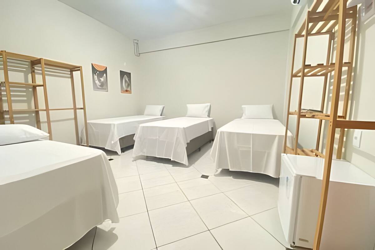 Une chambre d’hôtel avec trois lits simples, une tête de lit en bois, un sol carrelé et un mur blanc.