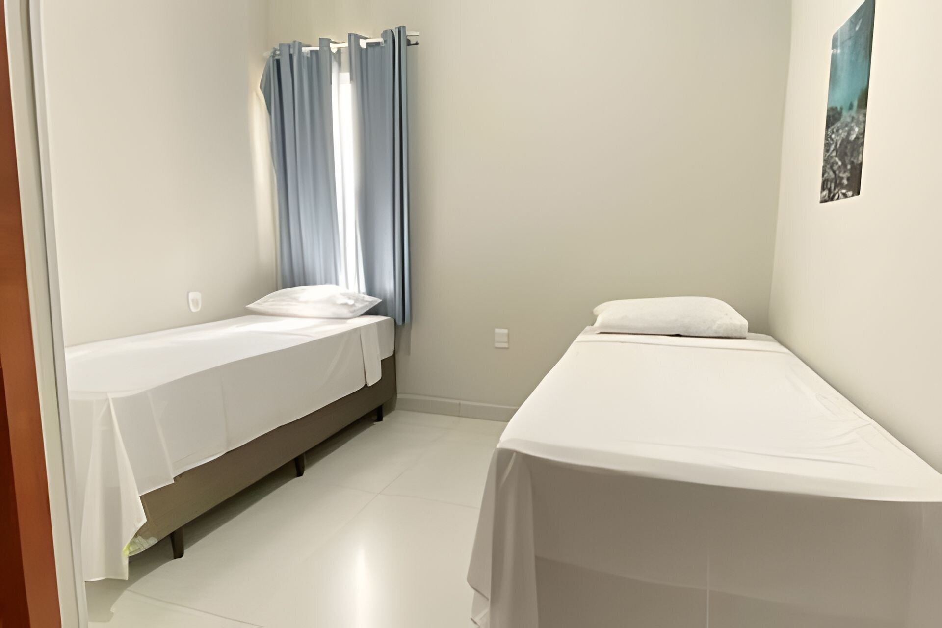 Quarto pequeno e limpo, com duas camas de solteiro, janela com cortinas e parede lisa.