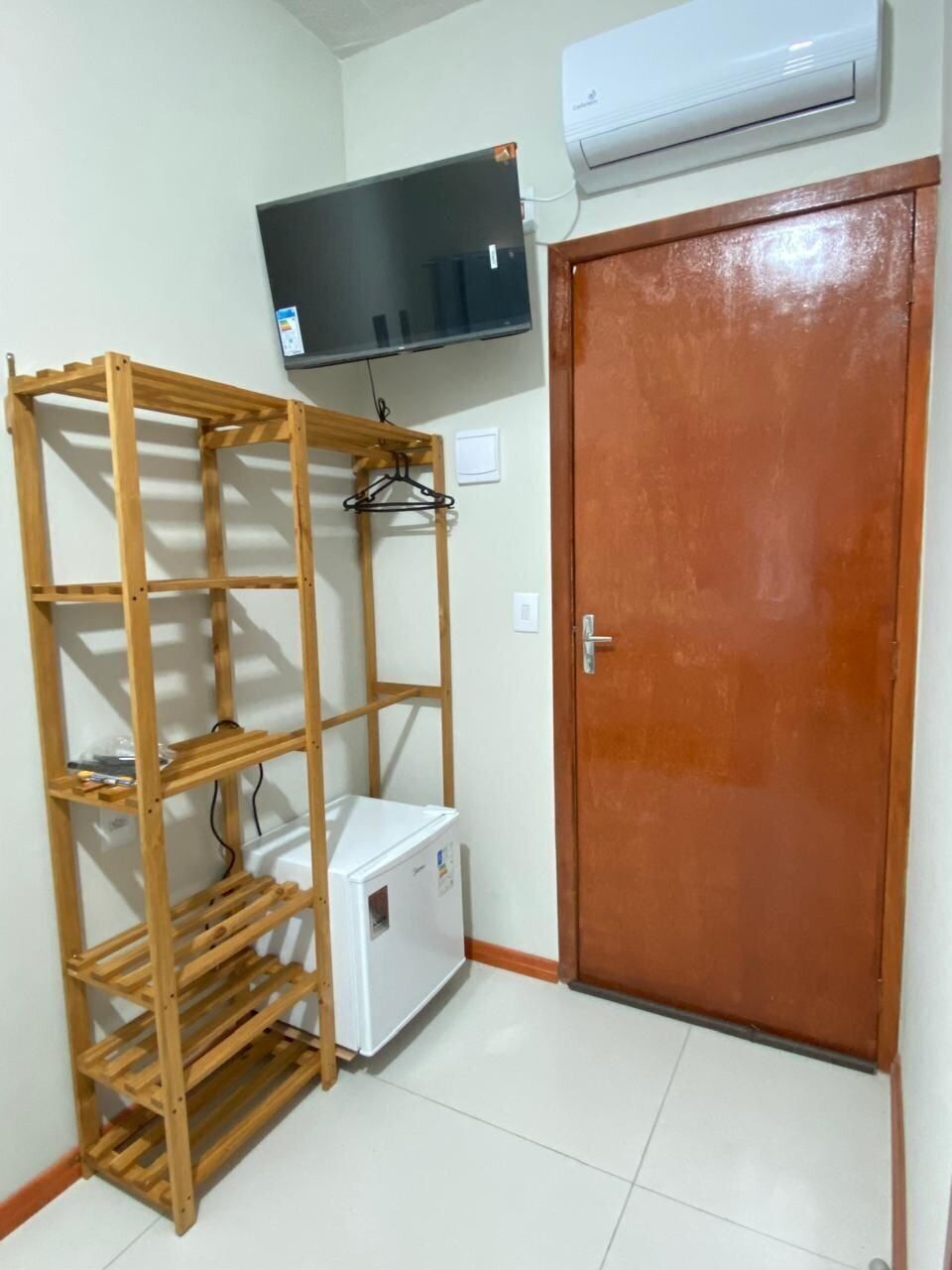 A room with a wooden shelving unit, a white refrigerator, a door, and a mounted television.