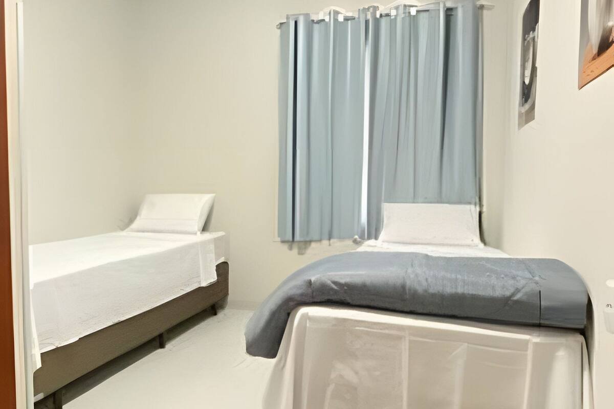 Une petite chambre propre, avec deux lits simples, une fenêtre avec des rideaux et un tableau accroché au mur.