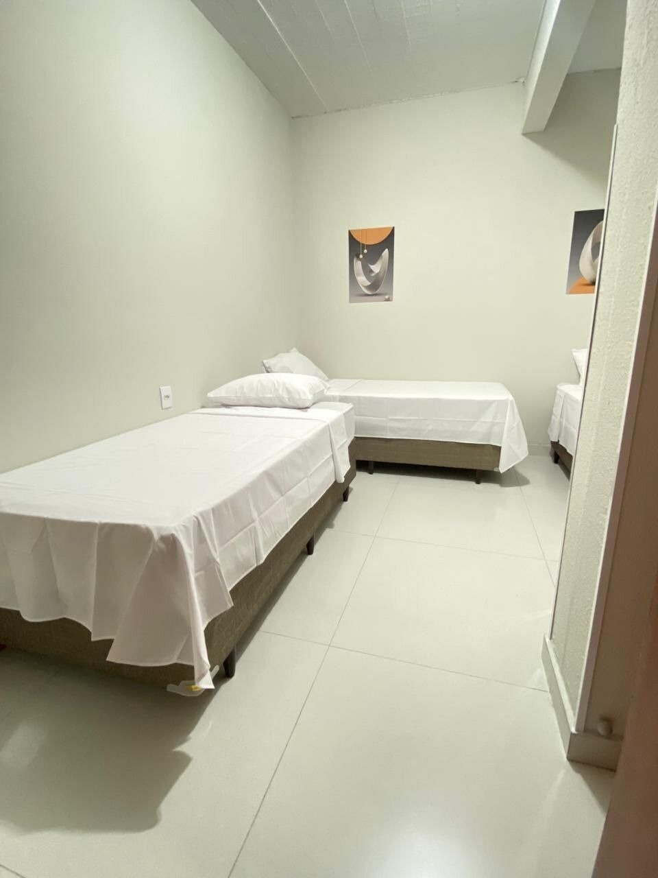 A small, clean room with two single beds, a white tiled floor, and a simple wall-mounted picture.