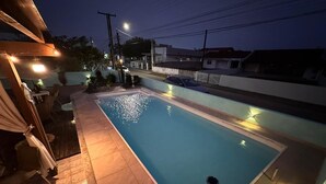 A rectangular swimming pool with a tiled deck, surrounded by a low wall and a pergola with string lights. - Casa praia da pinheira Piscina (Palhoça)