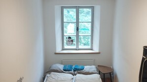 Une petite pièce étroite, avec un lit simple, une fenêtre donnant sur des arbres, un radiateur et une petite table.