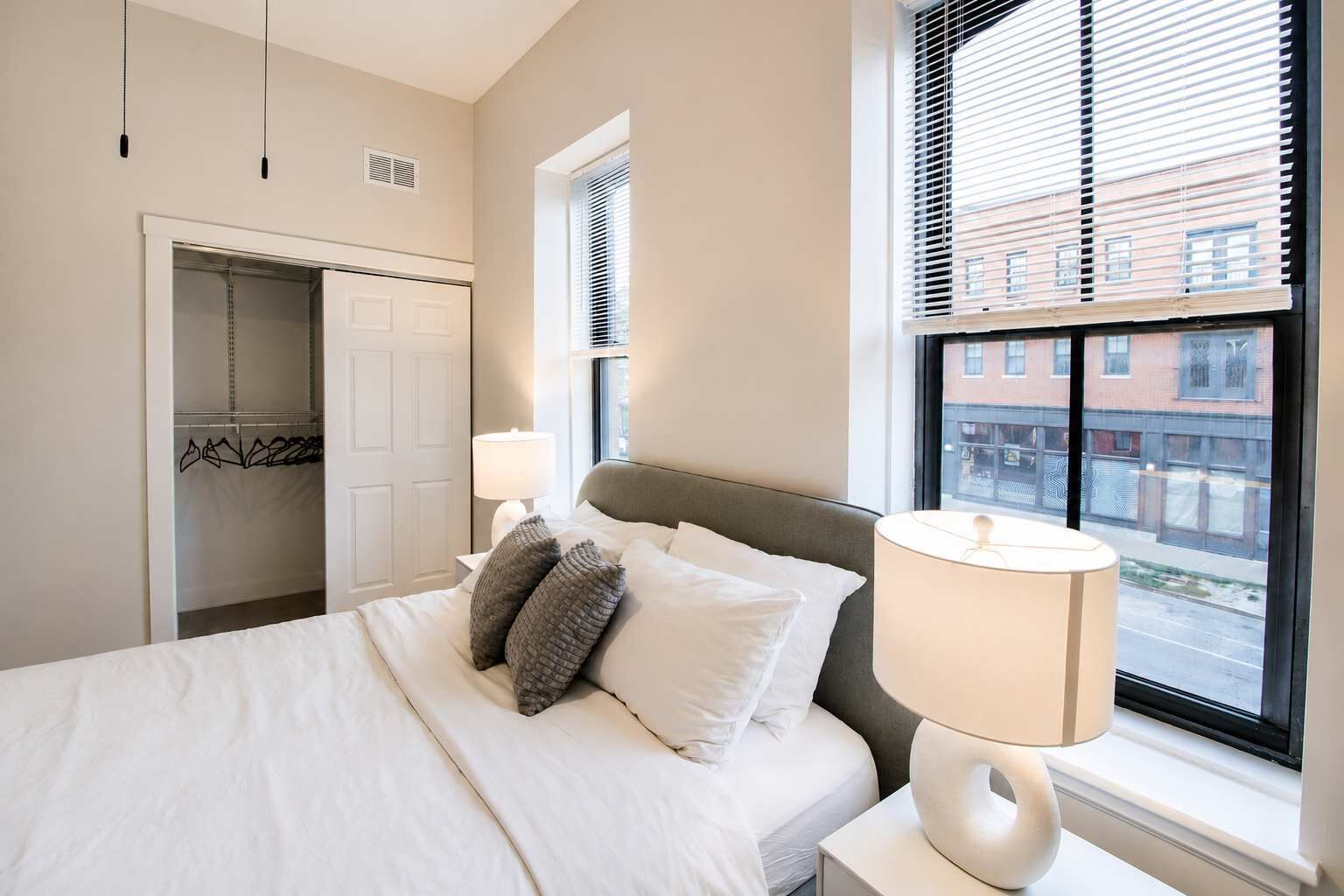 A bedroom with a bed, bedside lamps, a window with blinds, and a closet with hanging clothes.
