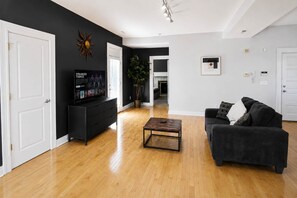 A living room with a black sofa, a coffee table, a TV on a black stand, and a plant. - 1BR Opposite SLU Hospital · Parking · Long Stays (St. Louis)