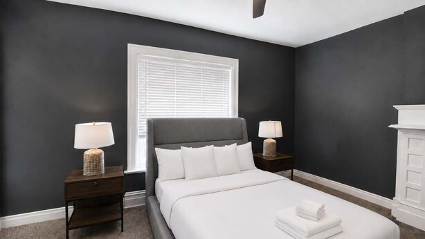 A bedroom with a bed, two bedside tables with lamps, a window with blinds, and a white dresser. - 1BR Opposite SLU Hospital · Parking · Long Stays (St. Louis)