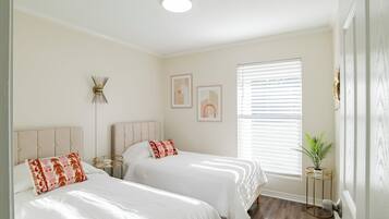 Un dormitorio con dos camas, una mesita de noche, una lámpara de pared, una obra de arte enmarcadas, una ventana con persianas y una planta en maceta.