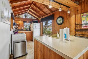 A kitchen with wooden walls, a refrigerator, a sink, and a clock. - Chipmunk Cottage | Cozy Lakefront Escape (Penn Yan)