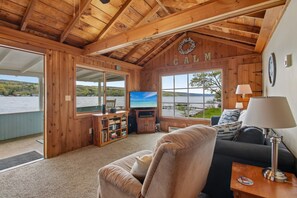A wooden cabin interior with a TV, sofa, and a view of a lake. - Chipmunk Cottage | Cozy Lakefront Escape (Penn Yan)
