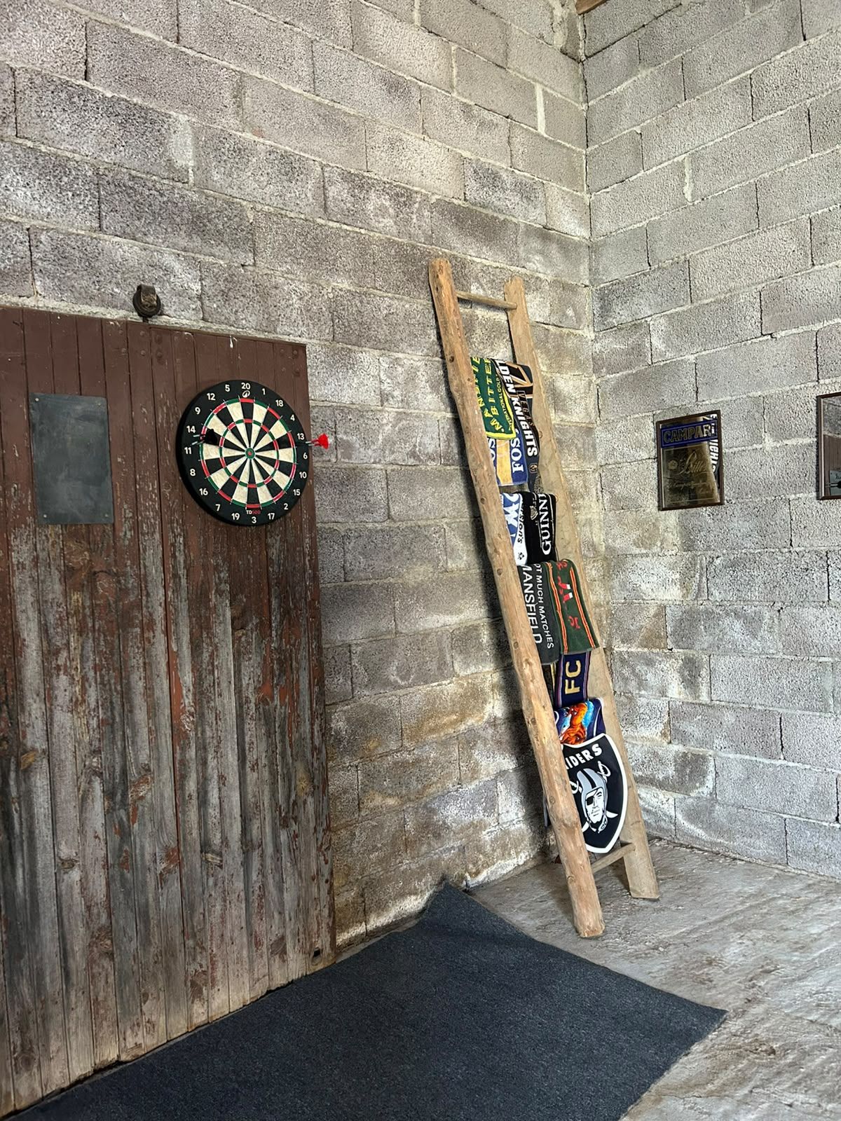 Een dartbord bevestigd aan een houten deur, een houten ladder rustend tegen een bakstenen muur, en een zwarte mat op de vloer.