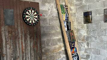 Een dartbord bevestigd aan een houten deur, een houten ladder rustend tegen een bakstenen muur, en een zwarte mat op de vloer.