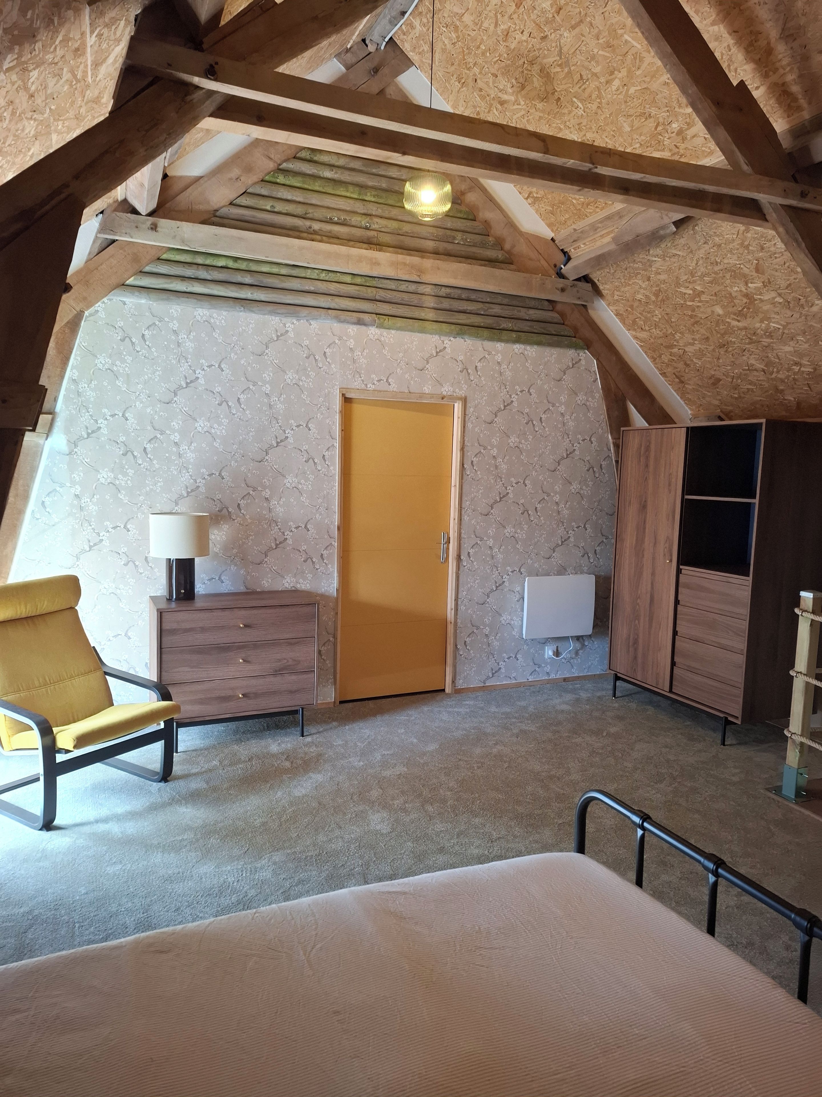 Ein Schlafzimmer im Dachgeschoss mit Holzbalkendecke, einem Bett, einem Sessel, einem Schminktisch und einem Kleiderschrank.