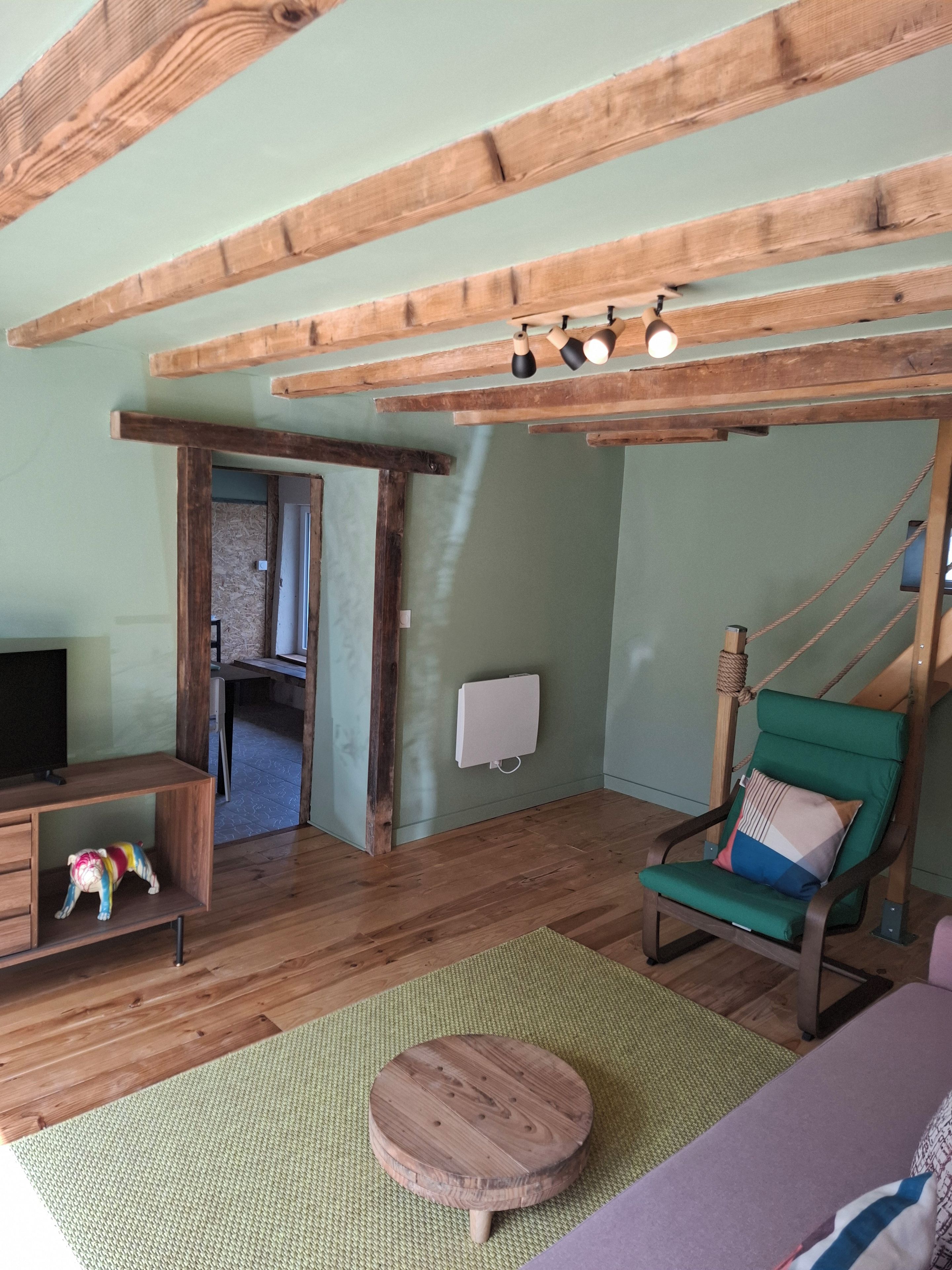 Ein Zimmer mit Holzbalken, einem grünen Sessel, einem Fernseher und einem kleinen Holzschemel.