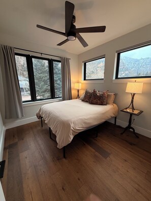 A bedroom with a bed, two bedside tables with lamps, a ceiling fan, large windows, and a view of mountains. - Beautiful 4-bedroom home in scenic Ouray, ideal for families and groups (Ouray)