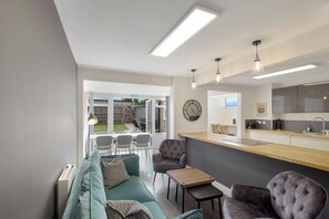 A modern living room with a sofa, chairs, a small table, and a kitchen area with a counter and cabinets. - 4BR Spacious Home | Garden & Parking | Long Term (Hertfordshire)