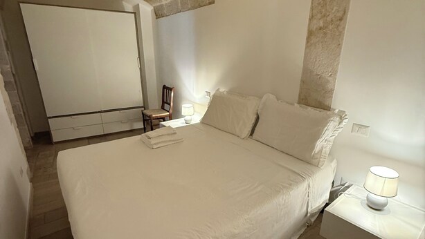 A bedroom with a stone ceiling, a bed with white bedding, a nightstand with a lamp, and a chair. - Mediterranean Deluxe Stone House (Molfetta)