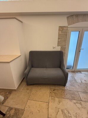 Un sofá gris en una habitación con suelo de piedra, pared blanca y otra pared de piedra con una puerta.