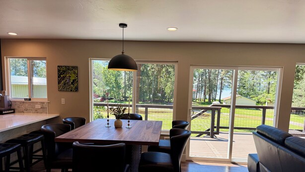Un espace repas avec une table en bois, des chaises noires, un luminaire suspendu et une vue sur une terrasse et la verdure à l’extérieur.