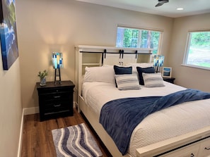 A bedroom with a bed, bedside tables, a nightstand, a window with a view of trees, and a ceiling fan.