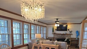 A spacious living room with a wooden dining table, chairs, a chandelier, a fireplace, and a television mounted on the wall.