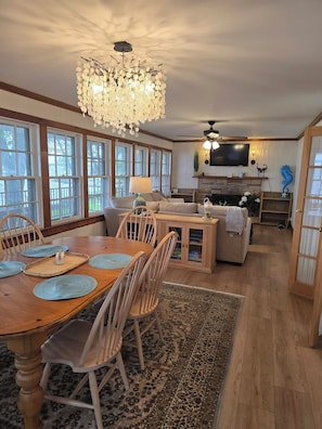 A spacious living room with a wooden dining table, chairs, a chandelier, a fireplace, and a television mounted on the wall.