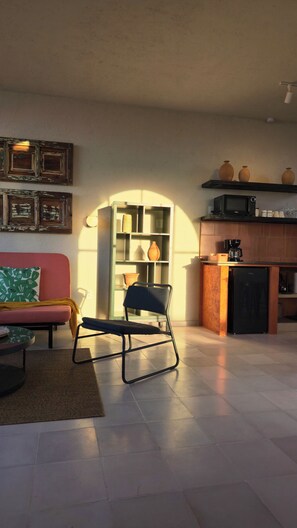 Ein Wohnzimmer mit einer Couch, einem Sessel, einem Couchtisch, einem Bücherregal und einer Küchenzeile mit Mikrowelle und Kaffeemaschine.