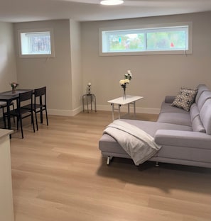 A living room with a grey sofa, a small white table with a vase of flowers, a dining area with a table and chairs, and two windows with views of greenery.